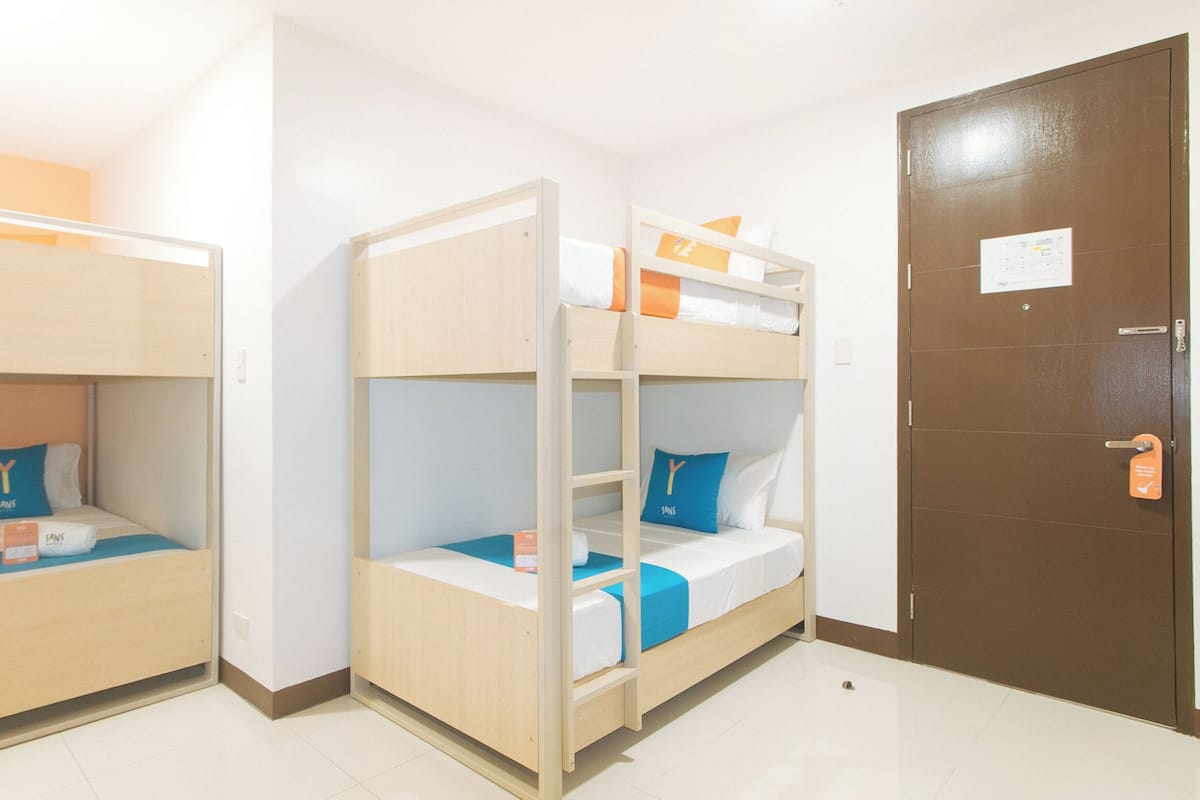A room with two bunk beds, each with a blue pillow and a white pillow with a letter 'Y'. There is a brown door with a sign on it, and a white wall with a light switch.
