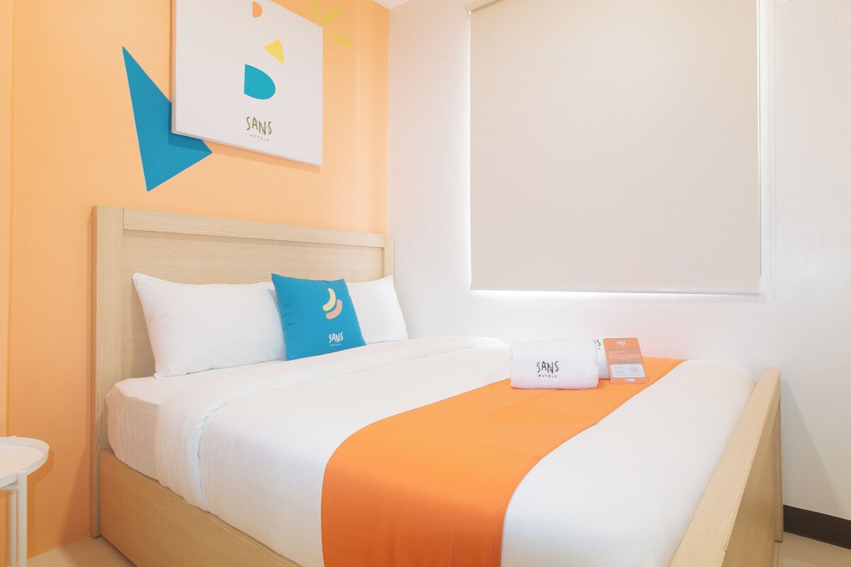 Habitació d'hotel amb un llit, una taula de nit, una obra d'art penjada a la paret i una finestra amb persianes.