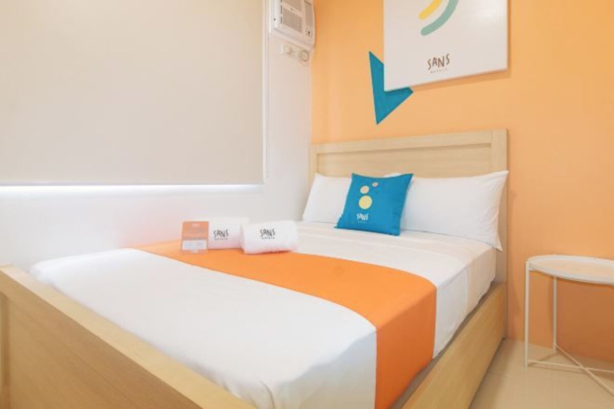 Una habitación de hotel con una cama, un edredón naranja, una almohada azul y una almohada blanca.