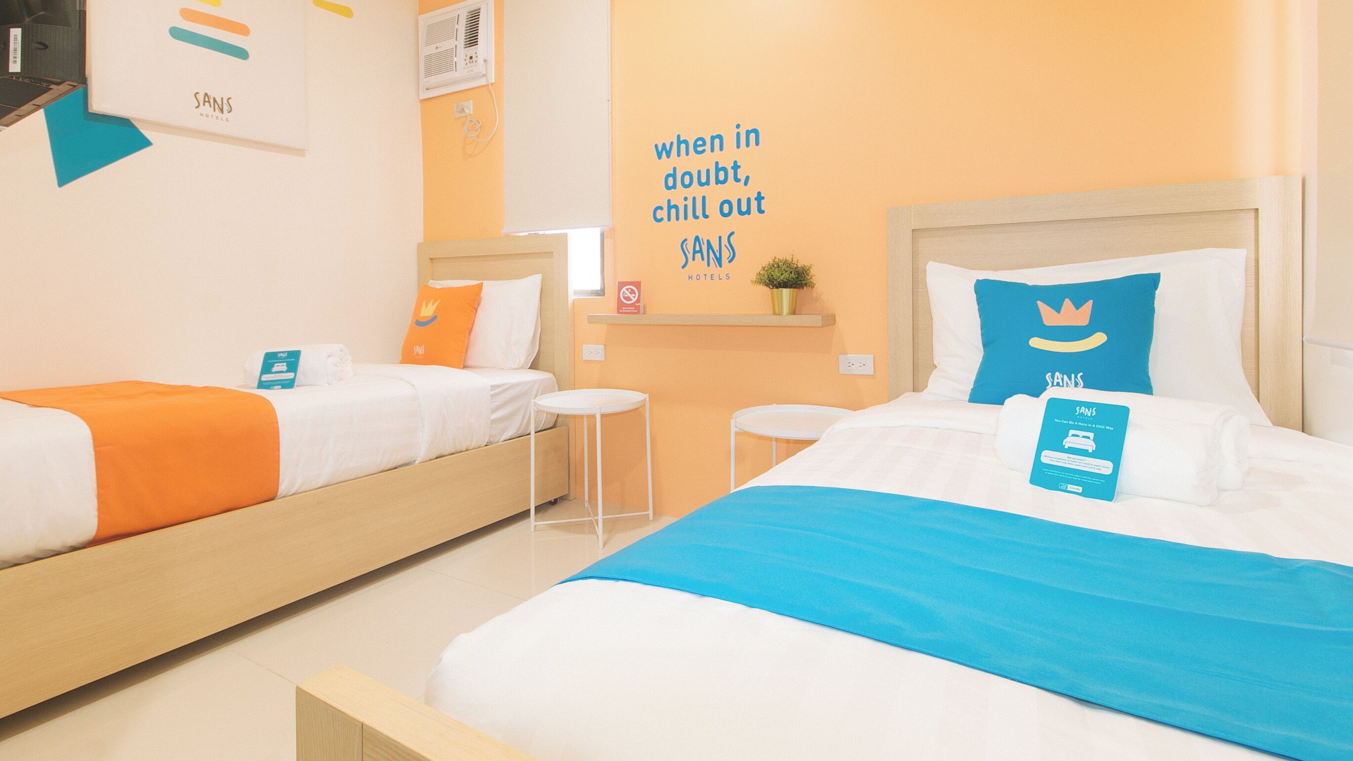 A hotel room with two beds, each with a different colored blanket, a small table, a chair, a wall-mounted shelf with a plant, and a motivational quote on the wall.