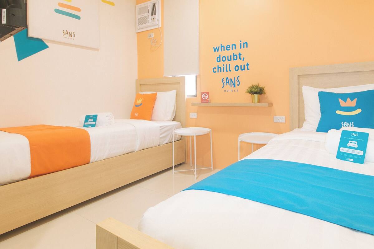 A hotel room with two beds, each with a different colored bedspread, a small table, a chair, a wall-mounted shelf with a plant, and a motivational quote on the wall.