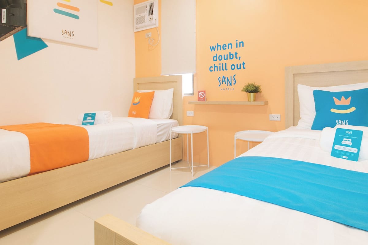 A hotel room with two beds, each with a different colored blanket, a small table, a chair, a wall-mounted shelf with a plant, and a motivational quote on the wall.