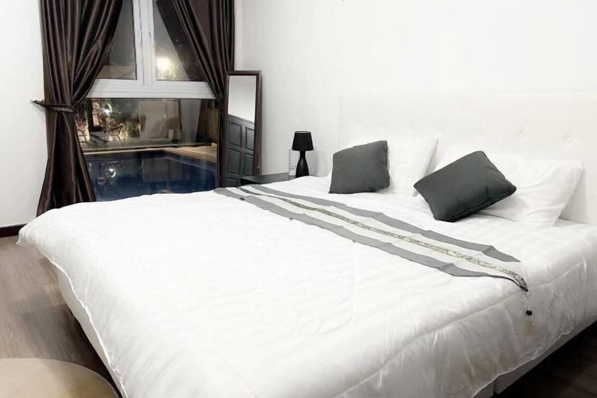 A neatly made bed with white bedding and two pillows, a bedside lamp, and a window with curtains.