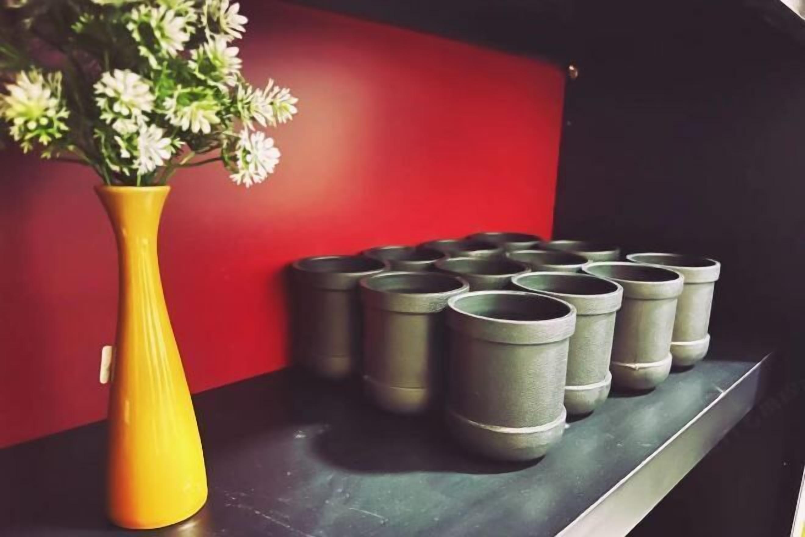 Un vase jaune contenant des fleurs blanches, posé sur une surface noire, à côté d’une rangée de jardinières grises adossées à un mur rouge.