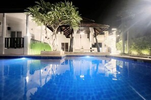 A swimming pool with a clear blue surface, surrounded by a modern building with white walls and a covered patio area.
