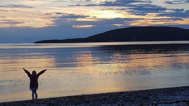 A person standing on a pebbled beach with arms raised, facing a calm body of water and a distant hill under a sunset sky. - Highland House in Ullapool (Ullapool)