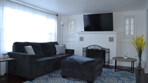 Uma sala de estar com um sofá cinza escuro, um banquinho combinado, uma lareira de tijolos brancos, uma TV de tela plana montada acima da lareira e uma pequena mesa redonda com um vaso de flores amarelas.