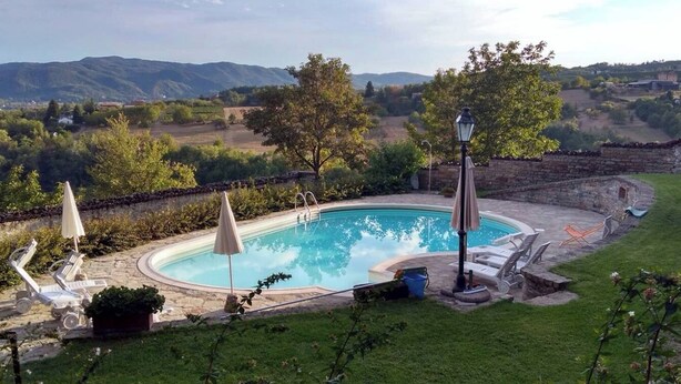 A swimming pool with lounge chairs and umbrellas, surrounded by a stone wall and a grassy area with trees and hills in the background. - Artemisia/bardana (Piemonte)