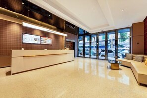 Een moderne hotelontvangst met een receptiebalie, digitale informatiepanelen en een zithoek met uitzicht op de straat.