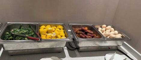 Un bufé con diversas opciones de comida, incluyendo verduras, maíz y salchichas.