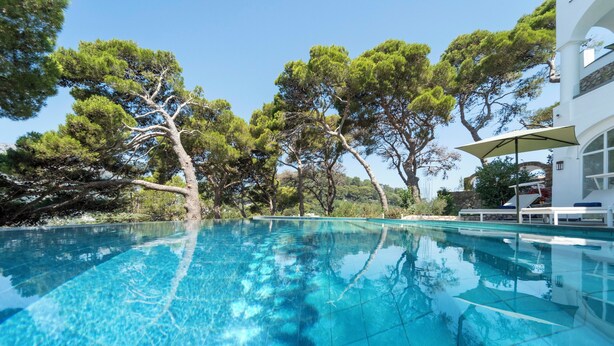 A clear blue swimming pool surrounded by trees, with a white building and outdoor seating area in the background. - Villa L'Ara (Capri)