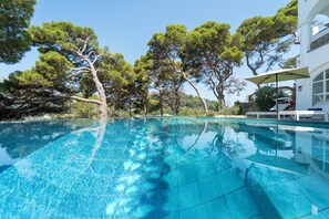 A clear blue swimming pool surrounded by trees, with a white building and outdoor seating area in the background. - Villa L'Ara (Capri)