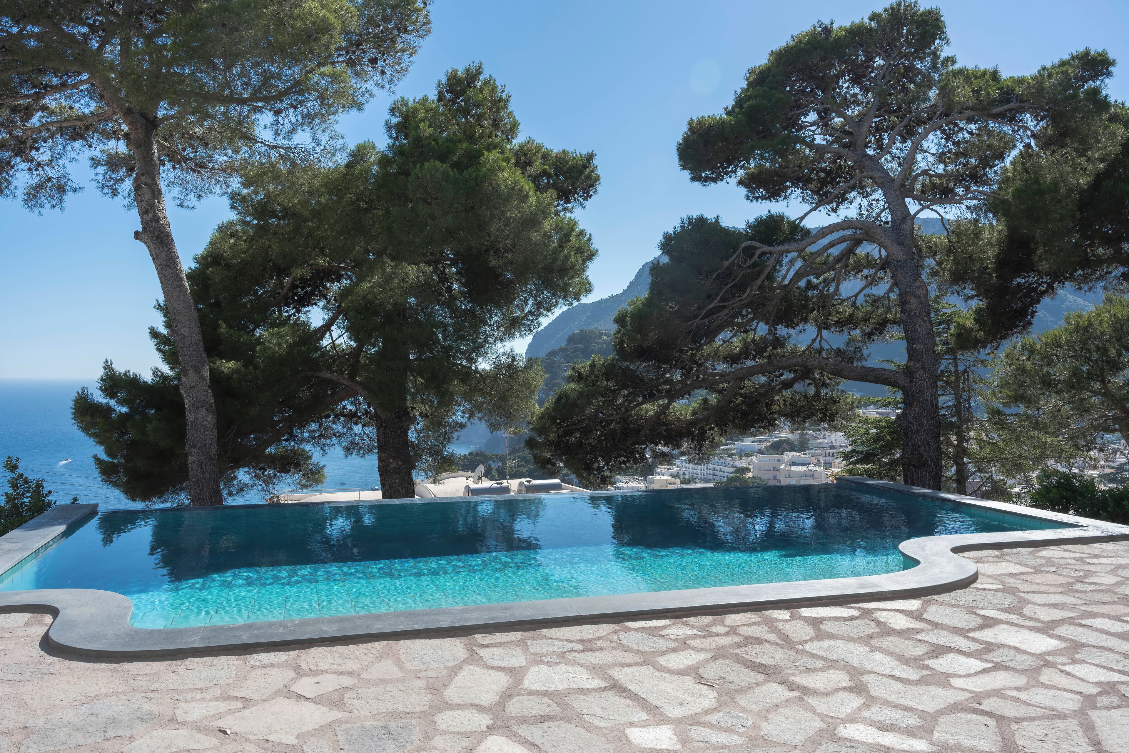 A swimming pool with a stone patio, surrounded by trees and overlooking a coastal landscape.