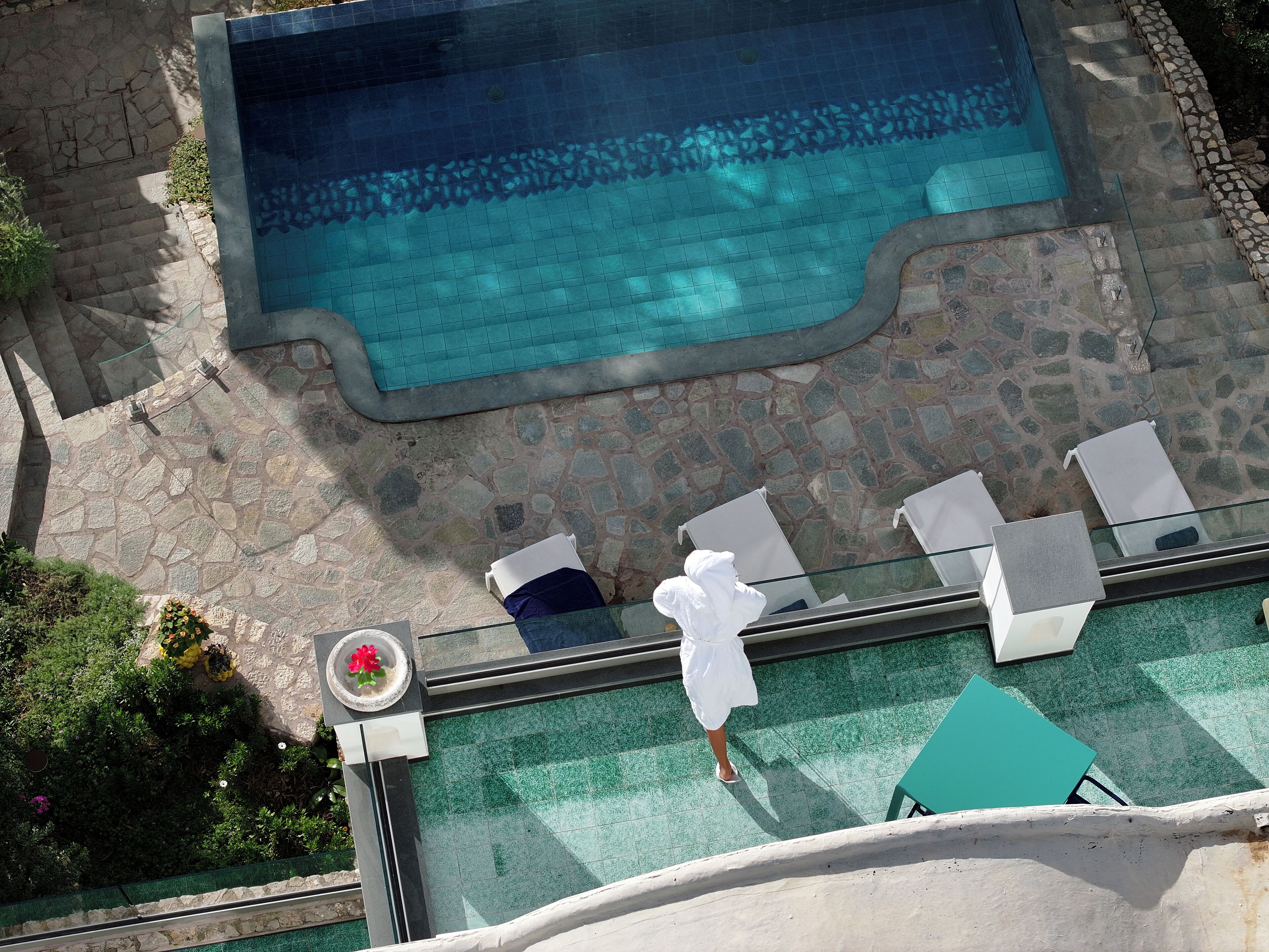 Luftaufnahme eines Schwimmbeckens mit Steinfliesen, Liegestühlen und einer Glasbrüstung.
