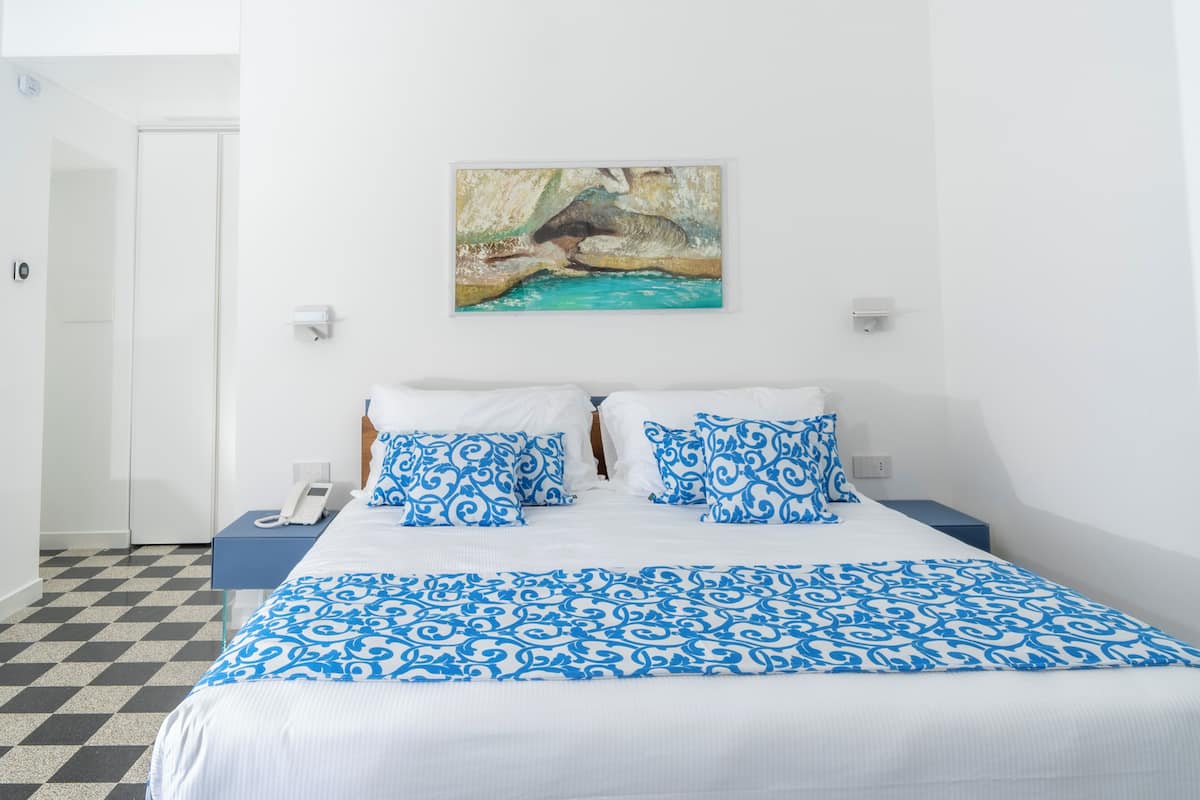 Une chambre d’hôtel avec un sol à carreaux, un lit avec une literie bleue et blanche, un tableau au mur et un téléphone sur la table de chevet.
