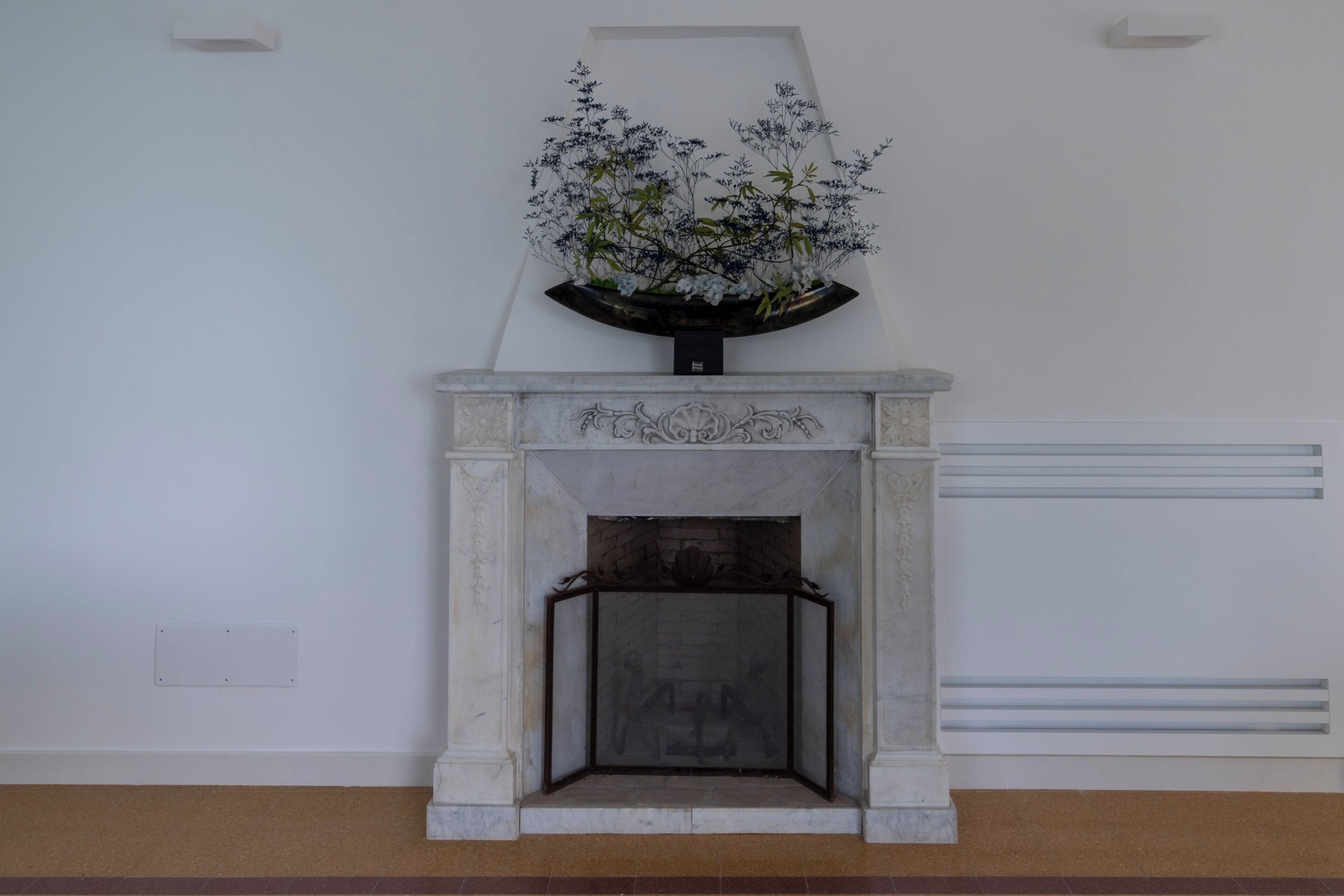 Een open haard met een decoratieve bloemstelling erboven, in een kamer met witte muren en een betegelde vloer.