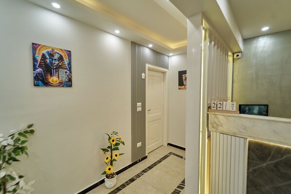 A hallway with a modern design, featuring a painting of an Egyptian figure, a potted plant, and a reception desk with a calendar.
