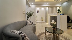 Ein modernes Wohnzimmer mit einem grauen Sofa, einem Glastisch und einer Kristallleuchter. Im Hintergrund ist ein Empfangsbereich mit einer Pflanze zu sehen.