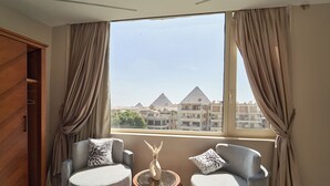 Ein Zimmer mit zwei grauen Sesseln, einem kleinen runden Tisch mit einer Vase und Blick auf Pyramiden durch ein großes Fenster.