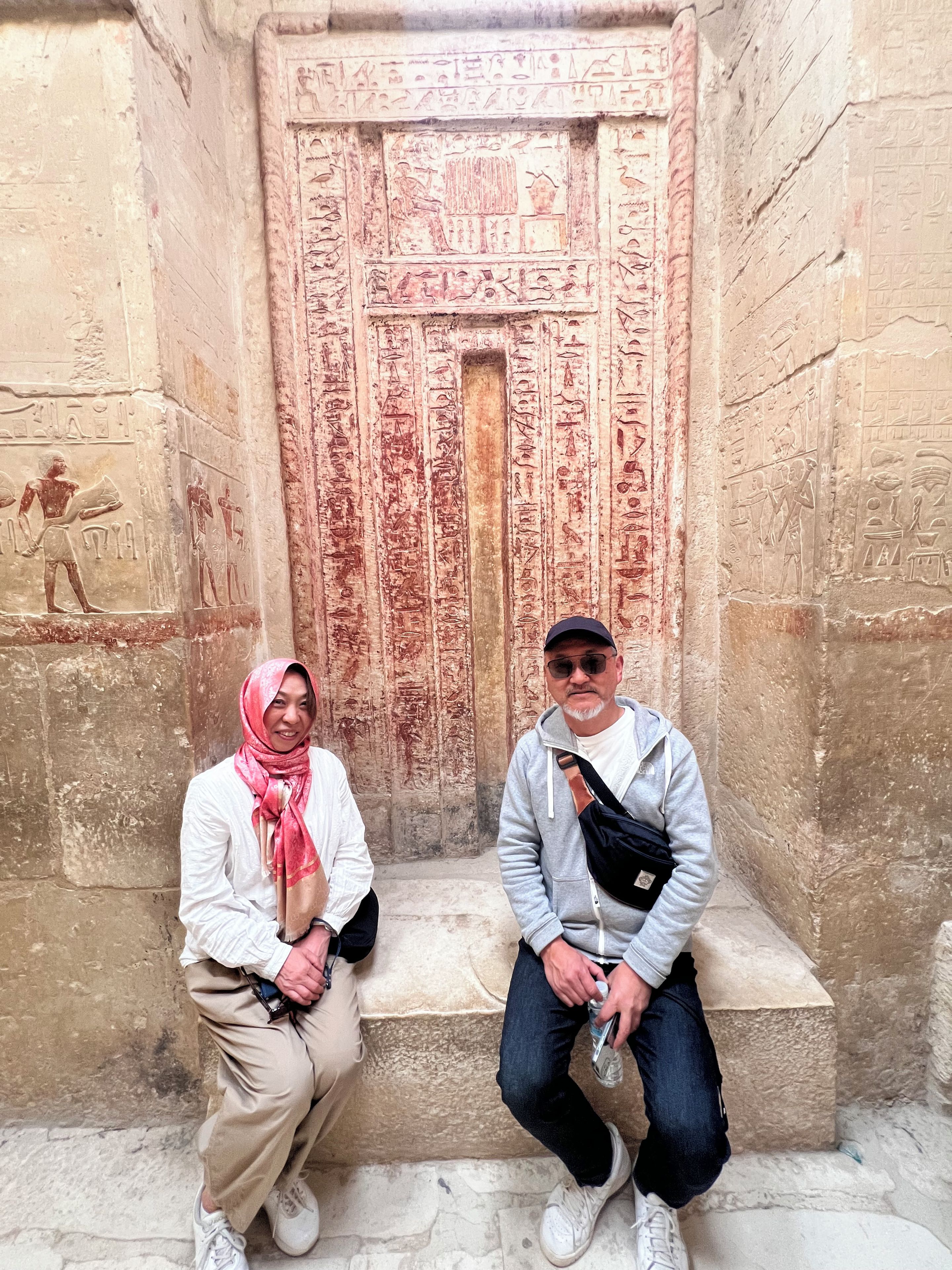 Deux personnes sont assises sur des marches devant un ancien mur égyptien orné de hiéroglyphes.