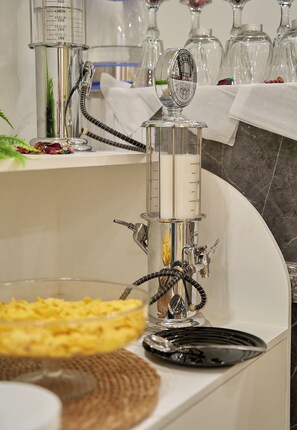 Ein Wasserpfeife mit transparentem Wasserbehälter und einer Messskala, platziert auf einer weißen Arbeitsplatte mit einer Glaskaraffe und einem Teller in der Nähe.