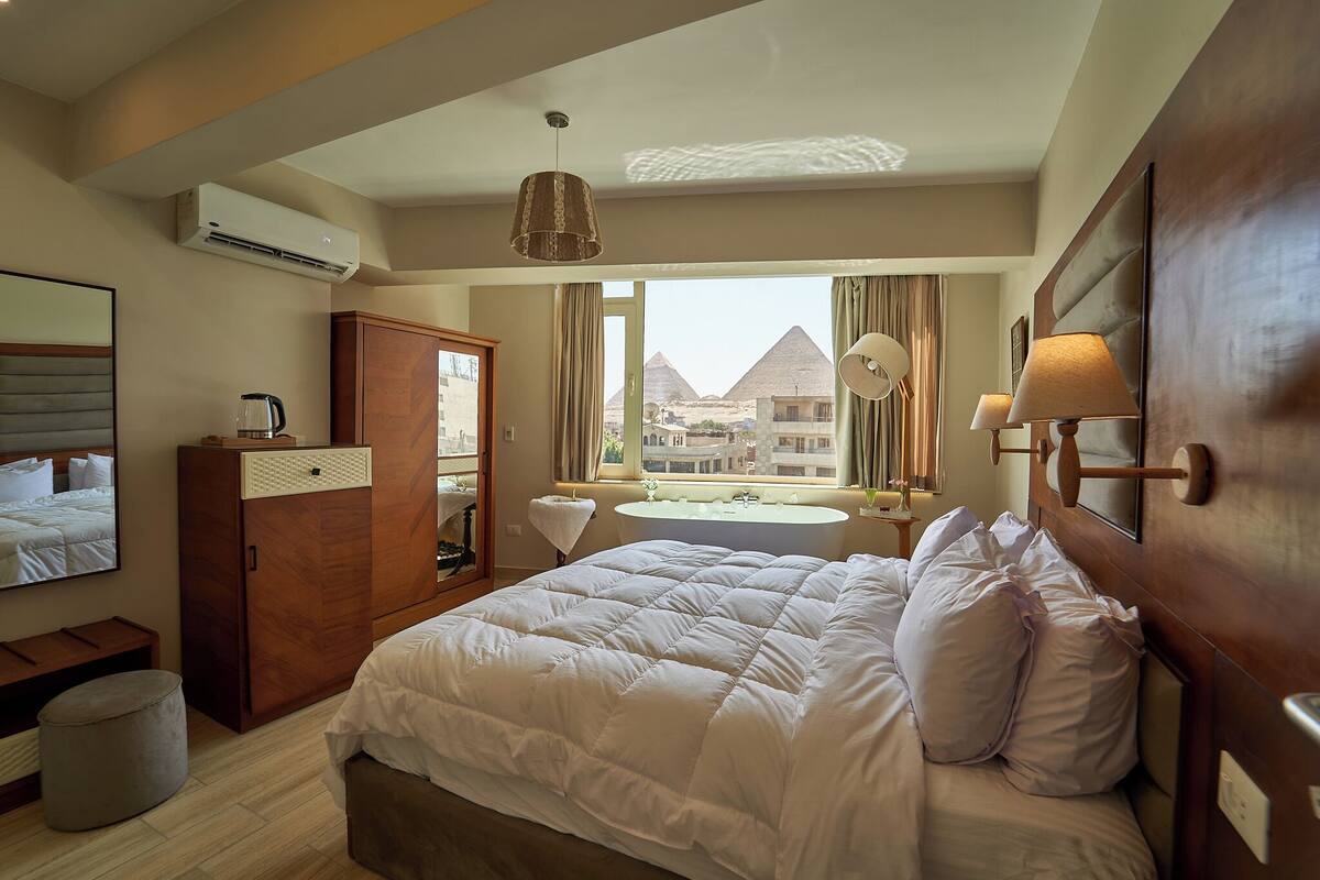 A hotel room with a large bed, a wooden wardrobe, a vanity with a mirror, a small table, and a view of the pyramids.