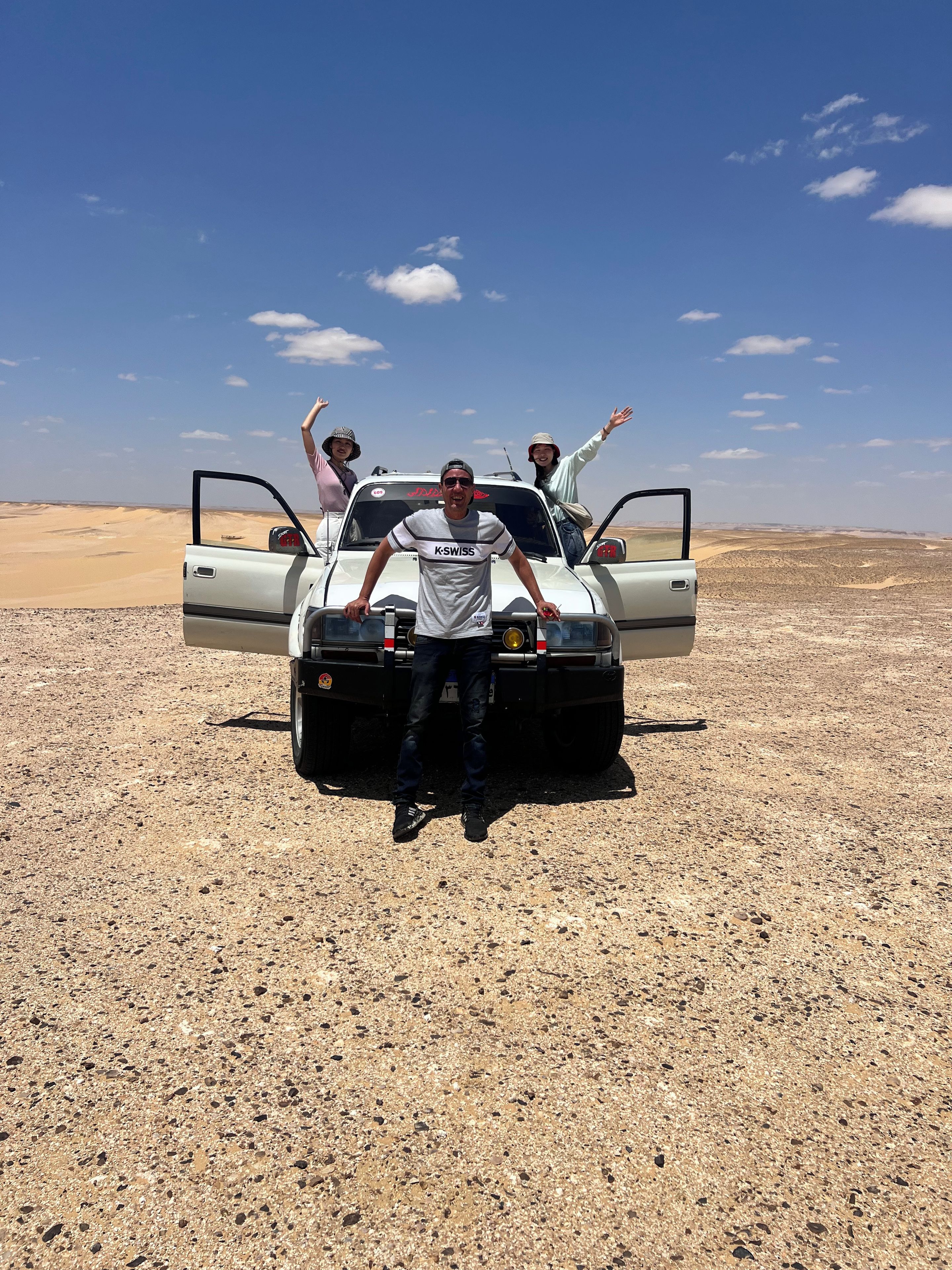 Wüstenlandschaft mit einem Fahrzeug und drei Personen davor.