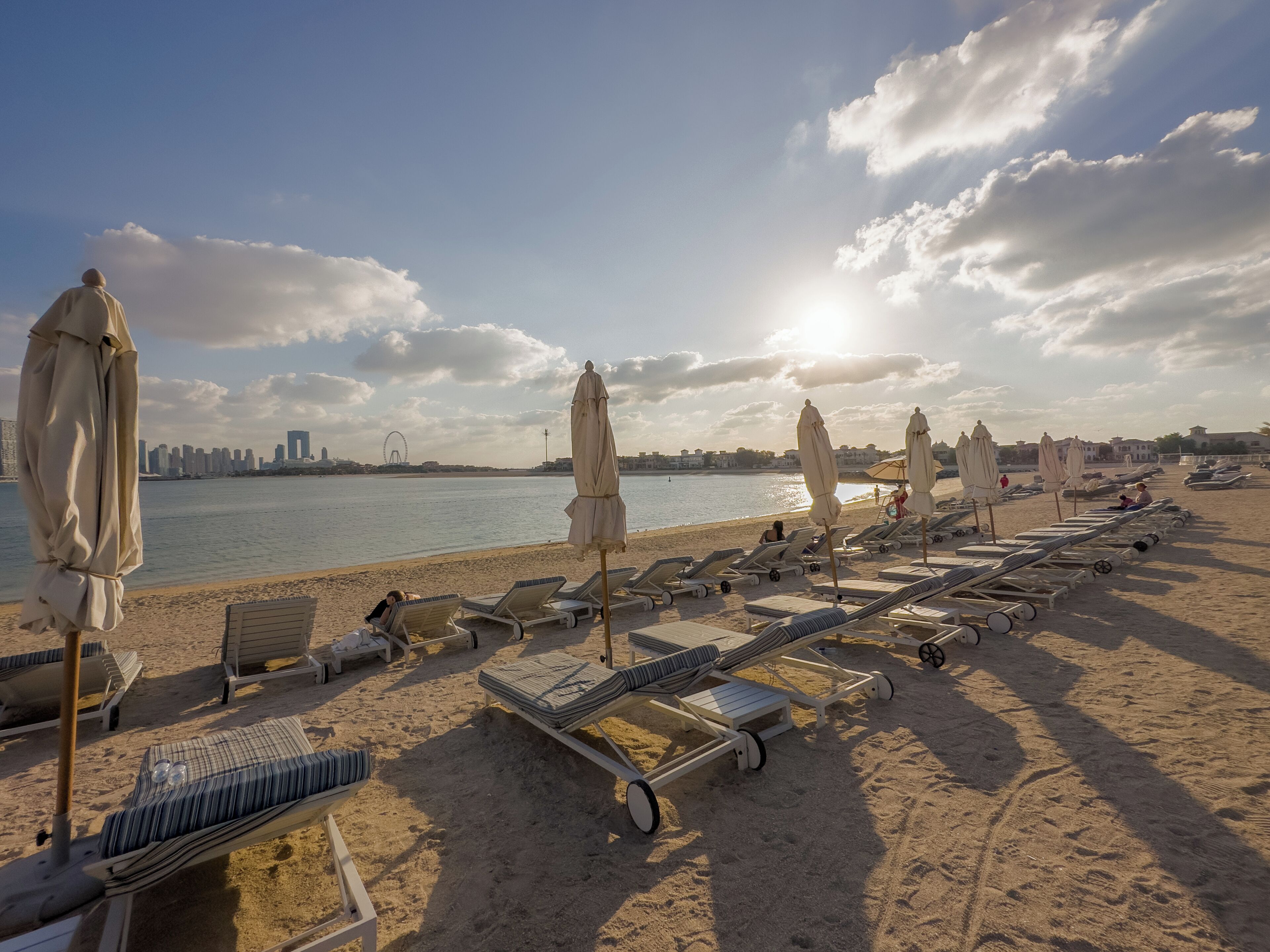 A beach with sun loungers and umbrellas, a clear sky, and a distant city skyline.