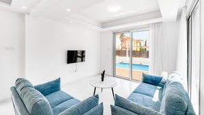 Ein modernes Wohnzimmer mit Flachbildfernseher, zwei blauen Sofas, einem kleinen weißen Tisch und einer großen Glasschiebetür, die zu einem Pool führt.