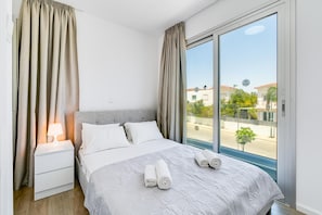 A hotel room with a large bed, a bedside table with a lamp, and a view of the street through a large window.