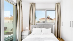 Ein modernes Schlafzimmer mit einem großen Bett, einem Nachttisch, einem Fenster mit Vorhängen und Blick auf Gebäude.