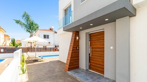 Ein modernes, zweistöckiges Haus mit Pool, Holztür und kleiner Außenbereich-Sitzgelegenheit.