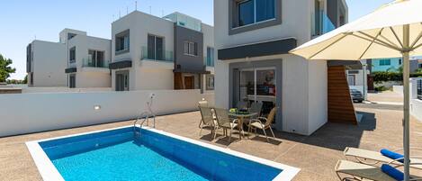 Un área de piscina moderna con una piscina rectangular, sillones de descanso y una mesa.