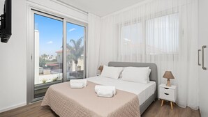 A bedroom with a bed, two bedside tables, a window with sheer curtains, and a view of buildings and palm trees outside.