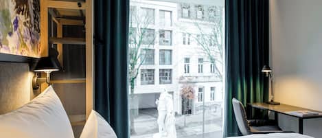 A hotel room with a bed, a desk, a chair, a statue, and a view of buildings outside.