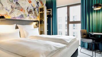 A hotel room with a large bed, a desk with a chair, a view of the city through a window, and a colorful abstract painting on the wall.