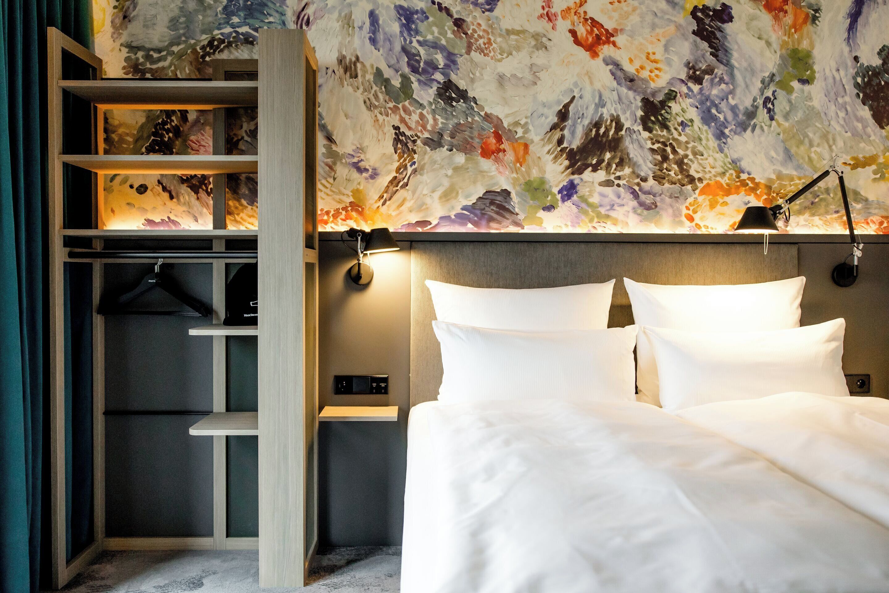 Habitación de hotel con cama, mesita de noche, lámpara, pared con un cuadro abstracto de colores y armario con repisas.
