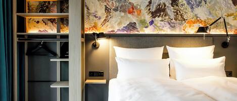 A hotel room with a bed, a nightstand, a lamp, a wall with a colorful abstract painting, and a wardrobe with shelves.