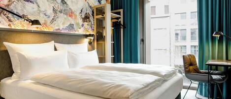 A hotel room with a large bed, a desk, a chair, a mirror, and a view of buildings through a window.
