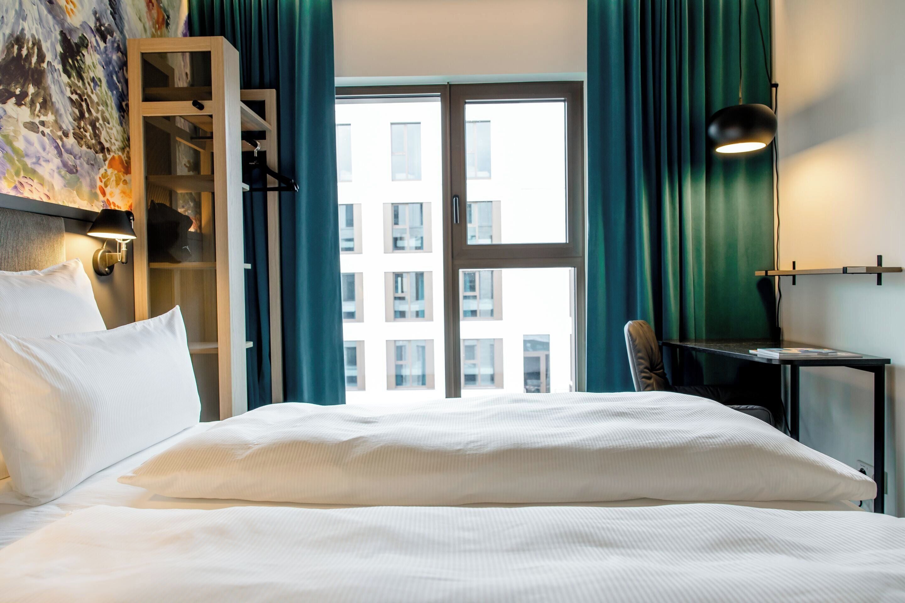 Quarto de hotel com cama, escrivaninha, cadeira, janela com cortinas e um quadro na parede.