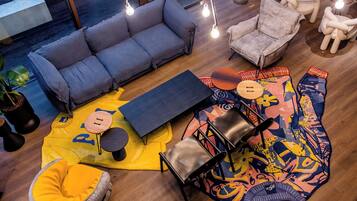 Een moderne woonkamer met een grijze bank, een geel vloerkleed en een kleurrijk abstract vloerkleed. Er staan twee stoelen en een klein tafeltje.
