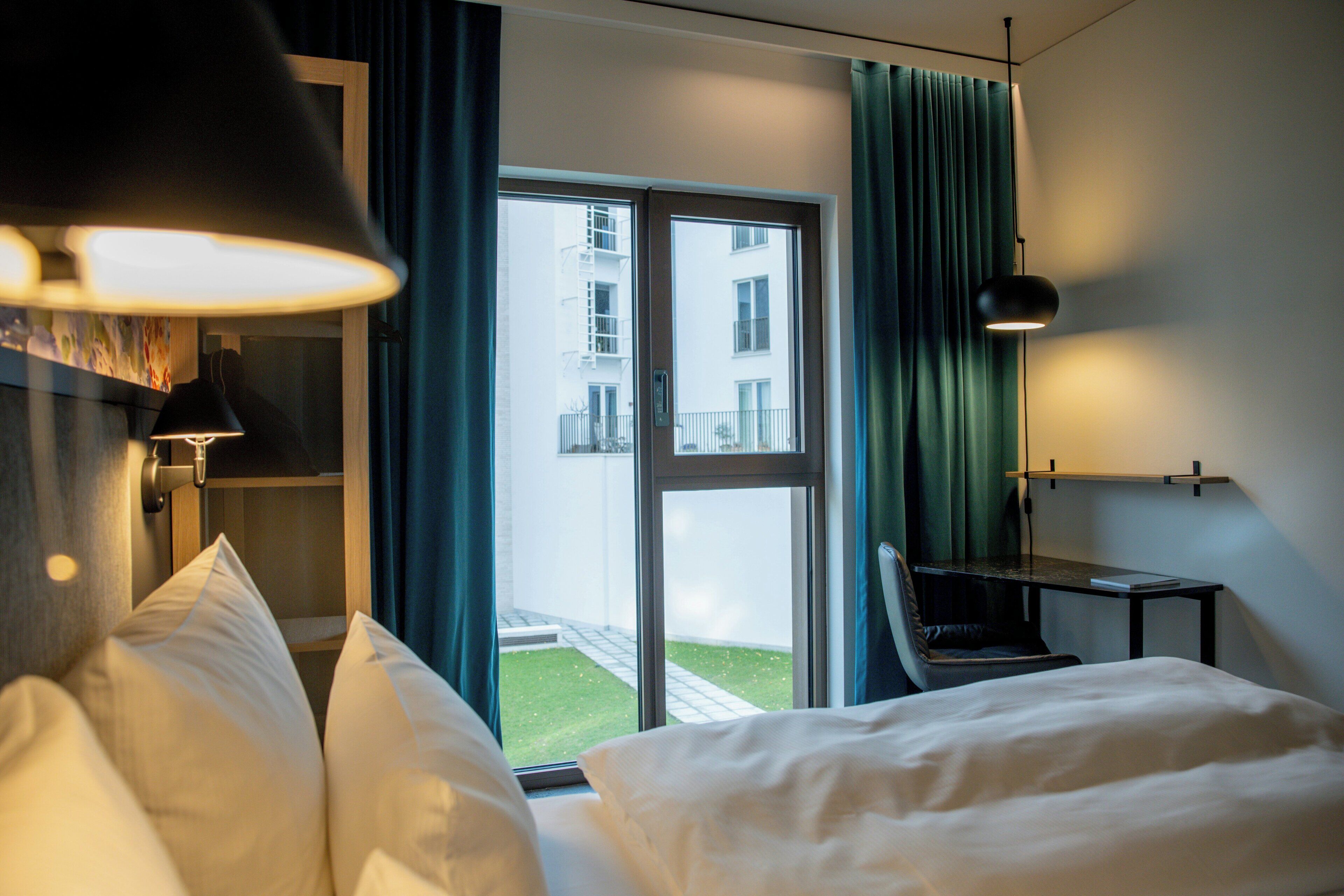 Habitación de hotel con cama, escritorio, silla, lámpara, ventana con cortinas y vista a un edificio.