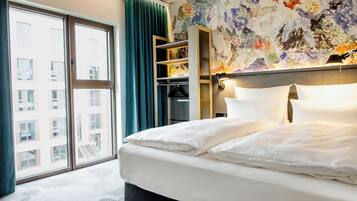 Habitación de hotel con una cama grande, un armario, un estante empotrado y vistas de la ciudad a través de un ventanal.