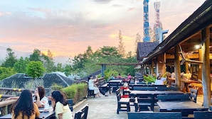 An outdoor seating area with wooden benches and tables, surrounded by greenery and a view of a telecommunications tower. - Khaopubpa Resort (Na Yong)