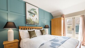A bedroom with a bed, two bedside lamps, a wooden nightstand, a large framed picture, and a wooden wardrobe. - Host Stay Heather Cottage (York)