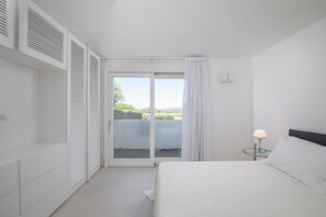 Un dormitorio con cama, mesita de noche, lámpara, puerta corrediza de vidrio a un balcón y un armario blanco.