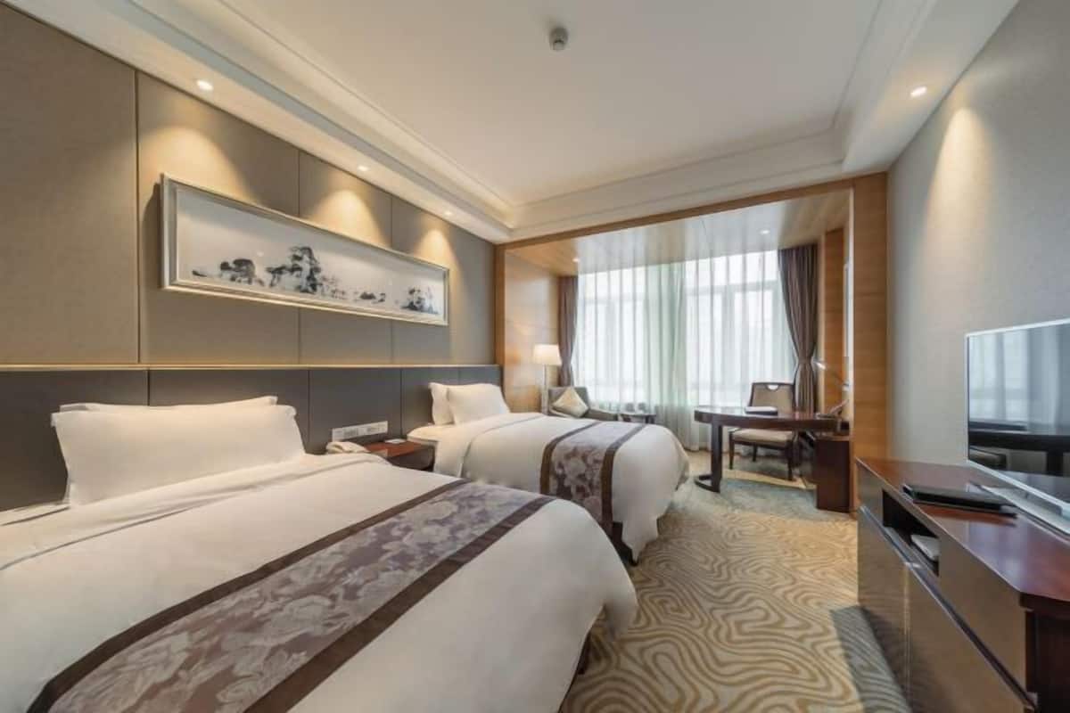 A hotel room with two beds, a desk, a chair, a TV, and a large window with curtains.