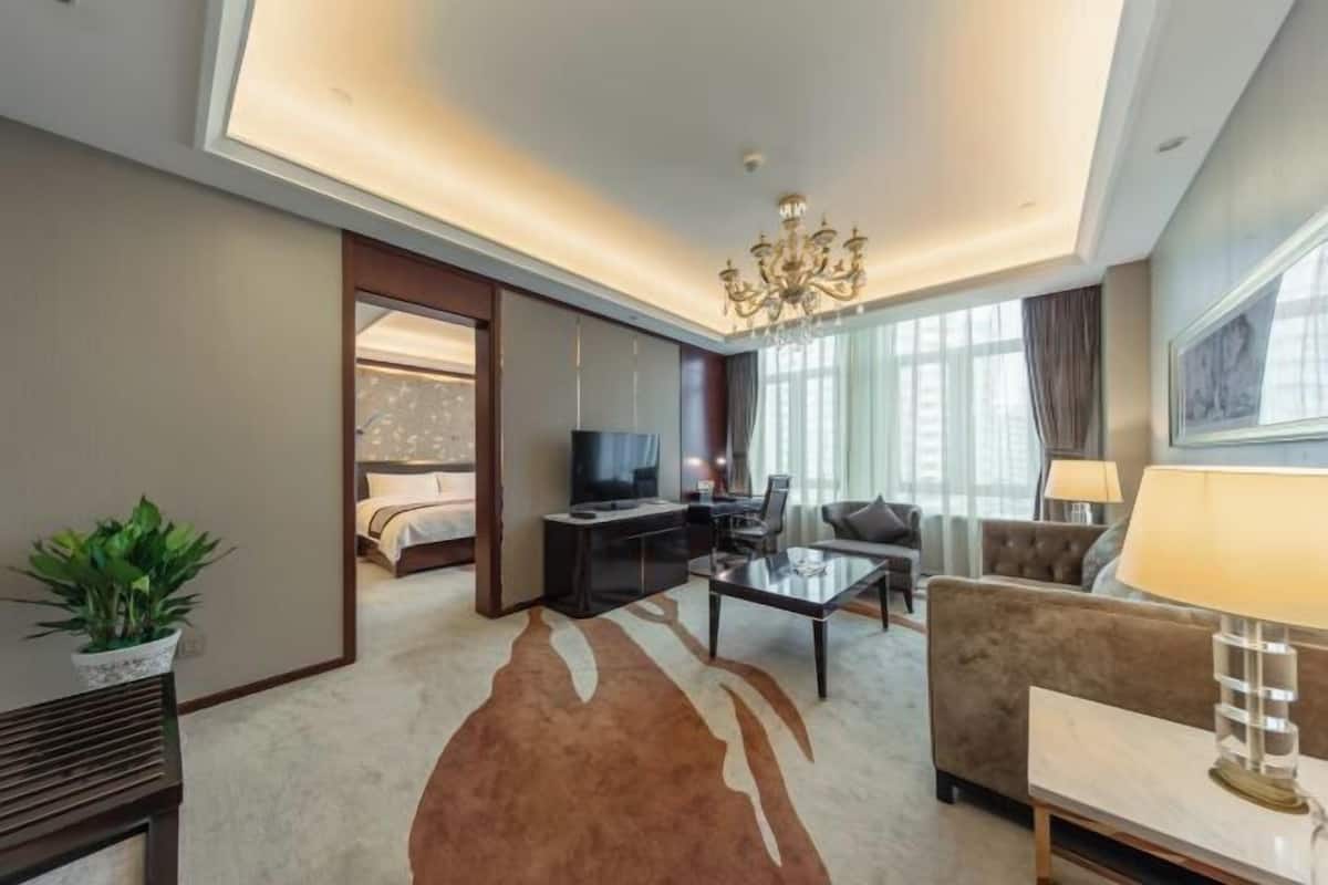 A hotel room with a living area, a television, a sofa, a coffee table, a chandelier, and a large window.