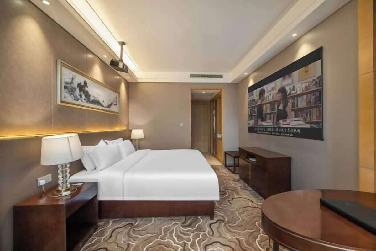 A hotel room with a bed, bedside tables, a desk, a chair, a wall-mounted TV, and a bookshelf.