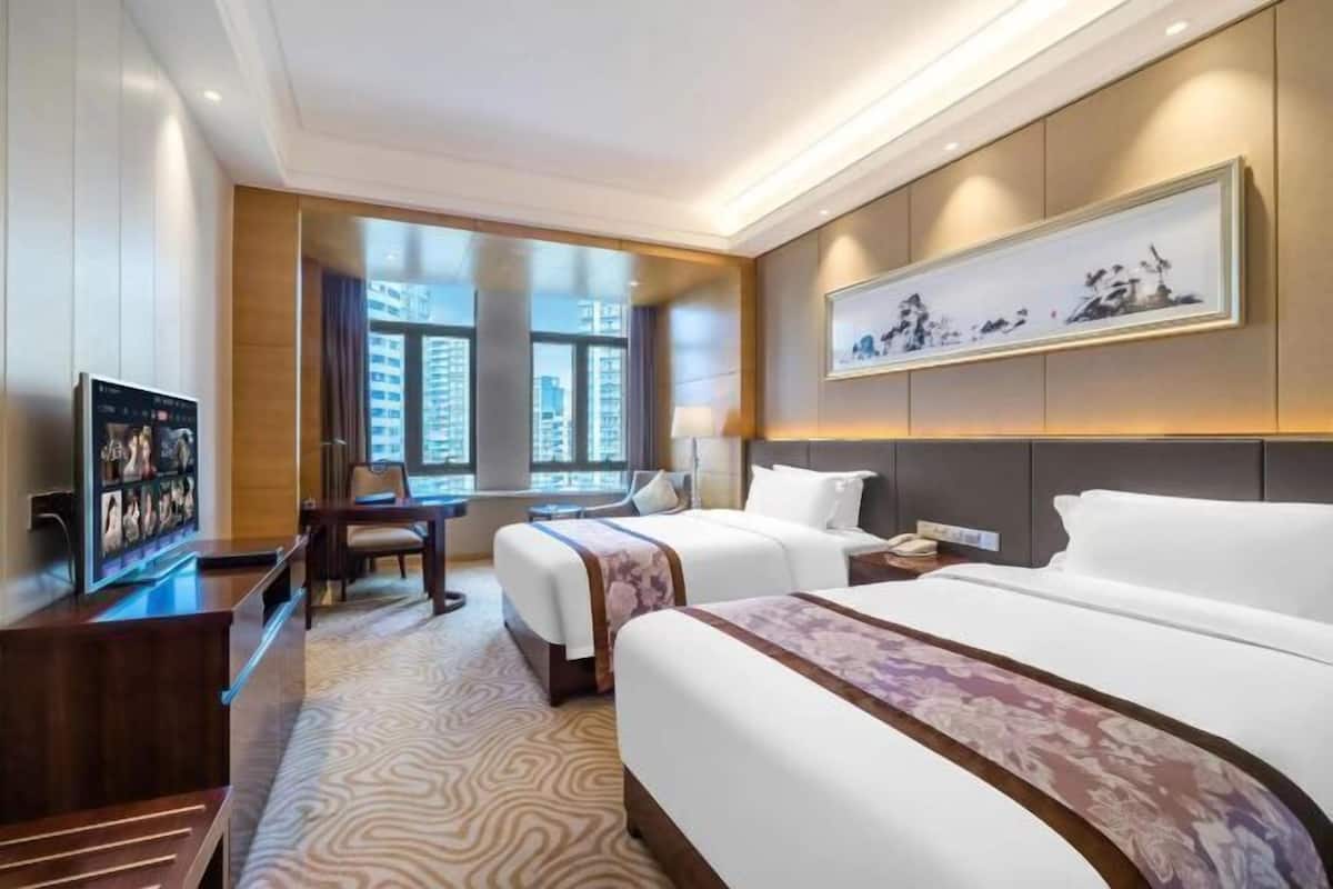 A hotel room with two beds, a desk, a TV, and a large window.